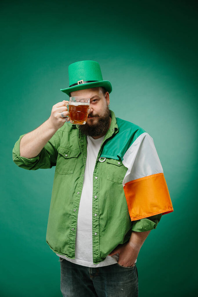 Bir bardak birası ve koyu yeşil arka planda İrlanda bayrağı olan mutlu sakallı bir adam. Aziz Patrick Günü. - Fotoğraf, Görsel