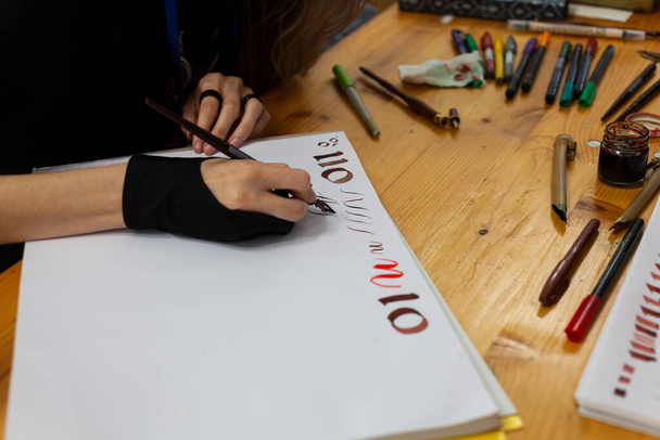 Erfahrene Kalligraphin übt sich in Schreiben und Zeichnen von Schriften im Renaissance-Stil in einem Skizzenbuch. Auf dem Holztisch liegen Marker und Buntstifte - Foto, Bild