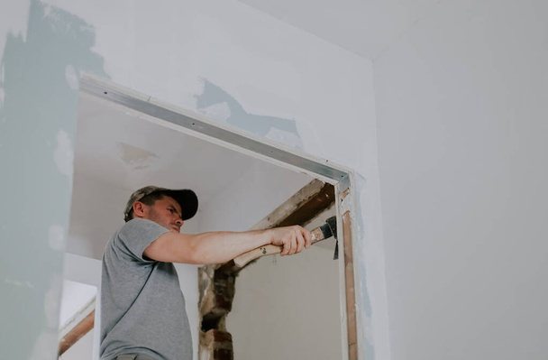 One young Caucasian man in a gray T-shirt and cap clears old putty from the beams of a doorway with one hand using an ax, close-up view from below with selective focus. Construction work concept. - Photo, Image