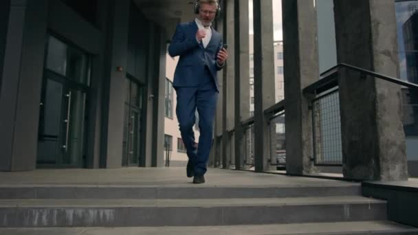 Activo divertido moviéndose bailando viejo hombre de negocios feliz de mediana edad caucásico maduro hombre de negocios caminar danza a la música escuchar canción en auriculares con auriculares y teléfono caminando por las escaleras oficina en la ciudad - Metraje, vídeo