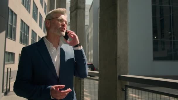 kaukaski prawnik inwestor pracodawca przedsiębiorca spacery na świeżym powietrzu stary dojrzały biały biznesmen smartfon negocjować telefon komórkowy rozmowy rozmowy starszy biznesmen chodzić w mieście w pobliżu biurowca - Materiał filmowy, wideo