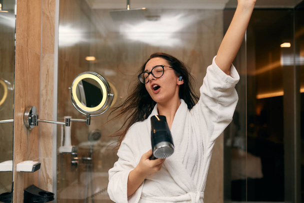 Radostná žena se sluchátky v uších si hravě osušuje vlasy fénem v koupelně po osvěžující koupeli, ztělesňující bezstarostný a energický moment sebepéče a relaxace.  - Fotografie, Obrázek
