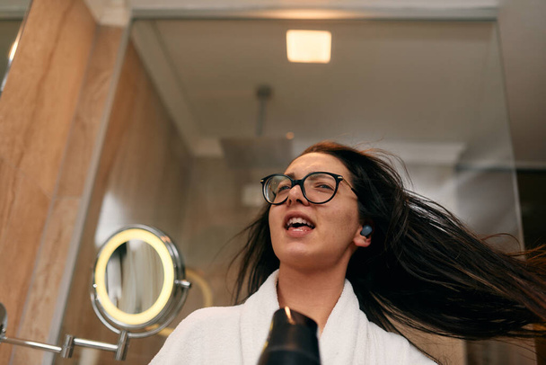 Радісна жінка, з навушниками у вухах, грайливо сушить волосся феном у ванній кімнаті після освіжаючої ванни, втілюючи безтурботний та енергійний момент самодопомоги та релаксації.  - Фото, зображення