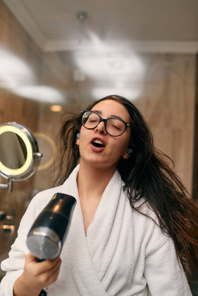 Iloinen nainen, jolla on kuulokkeet korvissaan, kuivuu leikkisästi hiuksiaan hiustenkuivaajalla kylpyhuoneessa virkistävän kylvyn jälkeen, jossa on huoleton ja energinen itsehoidon ja rentoutumisen hetki..  - Valokuva, kuva
