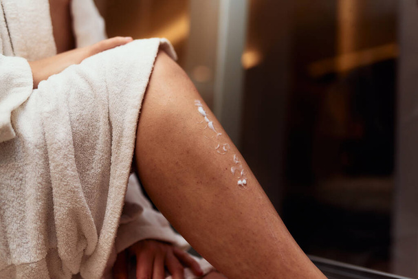 Een vrouw geniet van haar zelfverzorging routine, verwennen haar huid na een ontspannend bad door het aanbrengen van hydraterende crème op haar benen, omarmen de essentie van schoonheid en welzijn.  - Foto, afbeelding