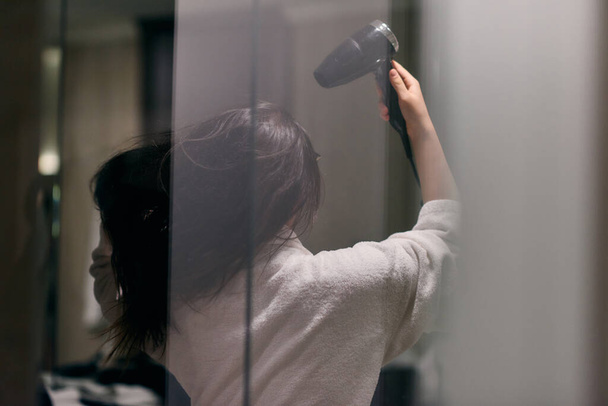 A joyful woman, with headphones in her ears, playfully dries her hair with a hairdryer in the bathroom after a refreshing bath, embodying a carefree and energetic moment of self-care and relaxation.  - Photo, Image