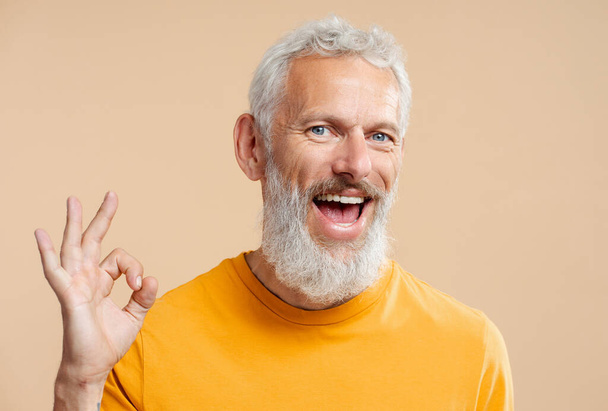 Komea hymyilevä 50-vuotias mies, jolla on tyylikkäät harmaat hiukset, yllään rento oranssi t-paita, joka näyttää ok, katsoen taustalla olevaa kameraa. Terveydenhuolto, hammashoito käsite  - Valokuva, kuva