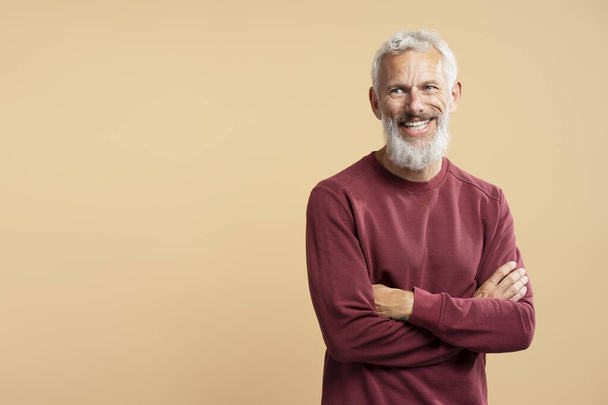 Портрет красивого улыбающегося бородатого мужчину, держащего скрещенные руки, смотрящего в сторону изолированного на бежевом фоне копировального пространства. Успешная бизнес-концепция  - Фото, изображение