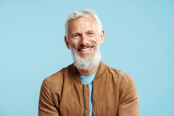 Bonito sorridente barbudo maduro homem com cabelo grisalho elegante vestindo jaqueta de couro marrom olhando para a câmera isolada no fundo azul  - Foto, Imagem