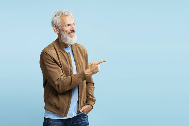 Handsome smiling gray haired European man pointing finger on empty space isolated on blue background. Advertisement, successful business concept  - Photo, Image