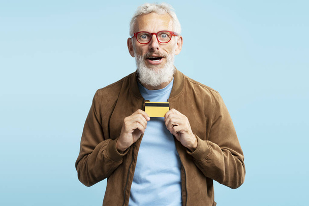 Retrato engraçado homem barbudo emocional segurando cartão de crédito dourado compras algo isolado no fundo azul. Dinheiro electrónico, conceito de cliente bancário  - Foto, Imagem