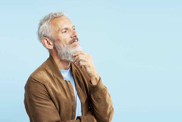 Portrait of pensive serious mature man touching his beard, planning project, brainstorming looking away isolated on blue background  - Photo, Image