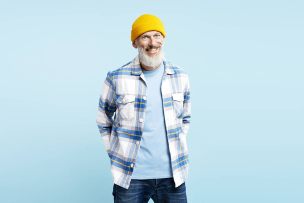 Portrét usmívající se atraktivní muž se žlutým kloboukem, stylové pruhované tričko izolované na modrém pozadí. Šťastný, sebevědomý turista, vousatý hipster hledící do kamery. Pozitivní životní styl, pojetí cestování  - Fotografie, Obrázek