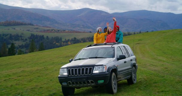 Grupa różnorodnych miłośników outdooru razem jeździć na wakacje w górach. Turyści stojący przez szyberdach samochodu śpiewają, tańczą i machają rękami na wycieczce. Turyści cieszyć jazdy w luku samochodowym. - Zdjęcie, obraz