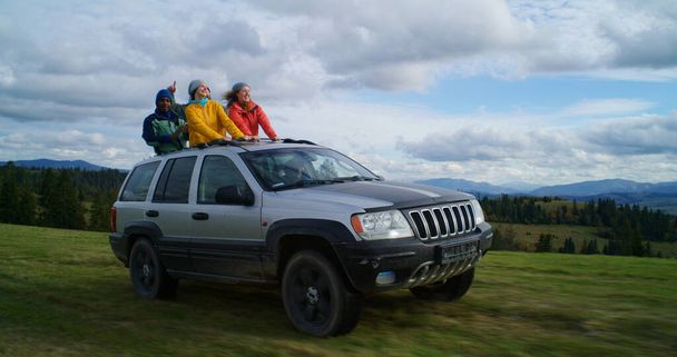 Grupa wielonarodowych turystów razem jeździć na wakacje w górach. Turyści stojący przez szyberdach samochodu, śmiejący się i machający rękami na wycieczce. Ludzie lubią jeździć w luku samochodowym. Miłośnicy pleneru - Zdjęcie, obraz
