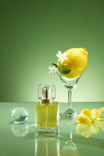 Scena reklamowa produktów zapachowych oleju cytrynowego. Prototyp butelki perfum nieoznakowany szklanym kubkiem zawierającym cytrynę i biały kwiat, kryształową kulę i skórkę cytryny na zielonym tle - Zdjęcie, obraz