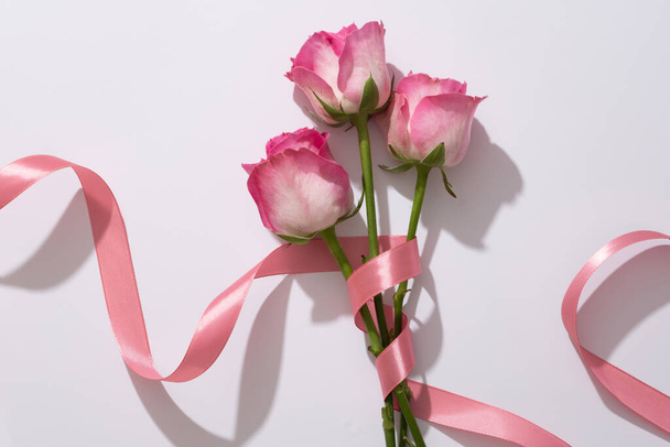 Zarte Rosen, die um ein weiches rosafarbenes Band auf weißem Hintergrund gewickelt sind. Flache Komposition mit schöner Blume für Werbung für Kosmetik- oder Designkarte, Postkarte und Banner - Foto, Bild