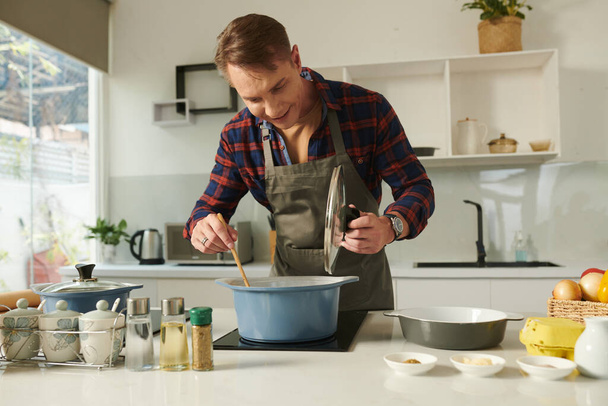 Hymyilevä mies sekoittamassa keittoa tai kastiketta saupanissa liedellä - Valokuva, kuva