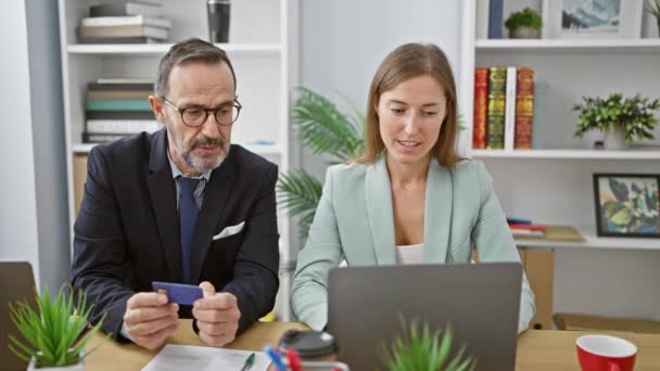 Δύο αξιόπιστοι επιχειρηματικοί συνεργάτες απολαμβάνουν ένα επιτυχημένο online shopping spree, χαμογελώντας μαζί καθώς χρησιμοποιούν το laptop και την πιστωτική τους κάρτα στο γραφείο - Πλάνα, βίντεο