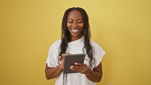 Una mujer sonriente con el pelo rizado sostiene una tableta delante de una pared amarilla lisa, que representa el uso casual de la tecnología. - Imágenes, Vídeo