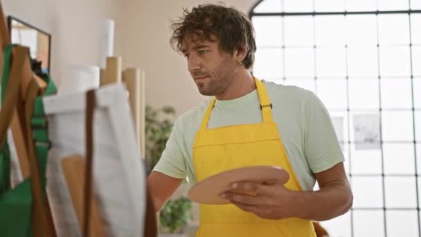 A smiling man in a yellow apron paints on a canvas in a bright art studio - Footage, Video