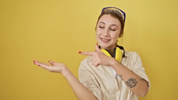 Amazed young brunette girl points with a confident smile, displaying safety glasses and noise reduction headphones over a yellow isolated background - Footage, Video