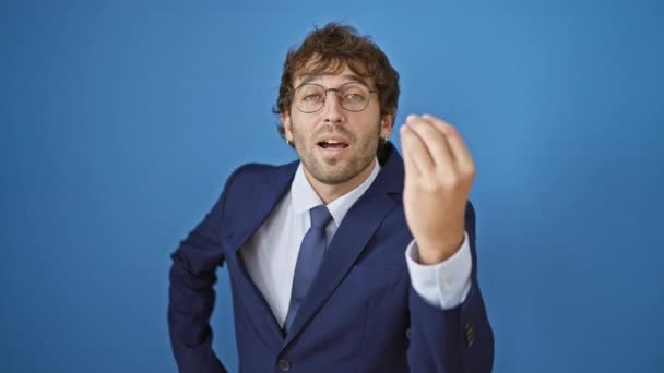 Уверенный в себе молодой человек в деловом костюме, осваивает итальянский жест пальцами, позируя на ярком изолированном синем фоне - Кадры, видео