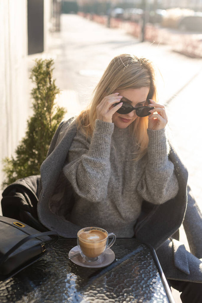 Giovane donna che si gode un caffè, seduta con il telefono cellulare sulla terrazza del caffè sulla vecchia strada della città durante una giornata di sole. Foto di alta qualità - Foto, immagini