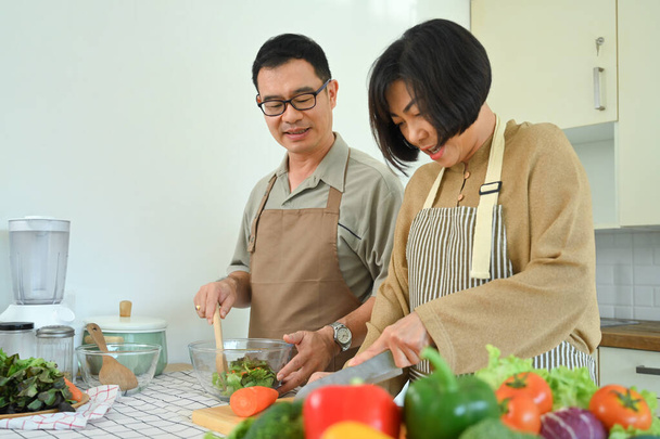 Ζευγάρι μέσης ηλικίας ετοιμάζει υγιεινό γεύμα ή δείπνο με φρέσκα βιολογικά λαχανικά στην κουζίνα. - Φωτογραφία, εικόνα