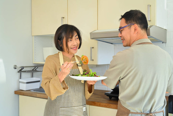 Ευτυχισμένο ζευγάρι Ασιατών ηλικιωμένων που φοράνε ποδιές και μαγειρεύουν μαζί στην κουζίνα του σπιτιού. Έννοια του τρόπου ζωής συνταξιοδότησης - Φωτογραφία, εικόνα