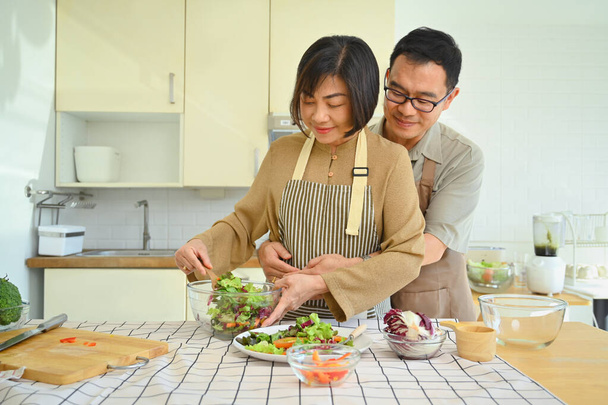 Loving middle age couple preparing healthy vegetables salad in kitchen. Happy senior lifestyle concept - Photo, Image