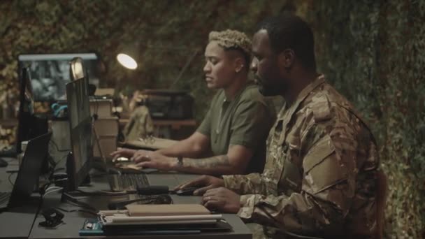 Średni spowolniony portret czarnego oficera wojskowego pozującego do kamery, siedzącego przy komputerze z widokami z kamer CCTV na ekranie, pracującego w wojskowym centrum kontroli z dwurasową koleżanką - Materiał filmowy, wideo