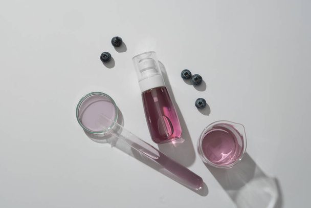 Üres spray palack, amely lila folyadékot, áfonyát és esszenciát tartalmaz fehér alapon díszített laboratóriumi üvegárukon. Mockup jelenet a reklámhoz. Természetes kozmetikai fogalom - Fotó, kép