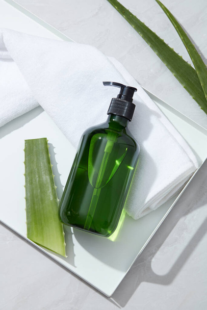 Top view of green plastic bottle unlabeled placed on white tray with towel and fresh aloe vera leaves on white background. Mockup for facial cleanser or shampoo product extracted from aloe vera. - Photo, Image