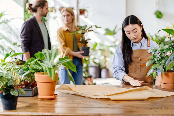 Charming Asian female worker in apron working behind counter, couple choosing plant in pot on blurred background. Plants business, start up. Small business concept. - Photo, Image