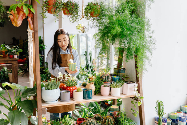 Onnistunut hymyilevä naispuolinen aasialainen kukkakauppias ja pienyrittäjä, joka nauttii työstään kasvien kanssa. Kukka suunnittelu studio, joten koristeet ja järjestelyt. Pienyrityksen käsite. - Valokuva, kuva