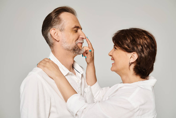 Portrait of middle aged smiling couple, woman touching man's nose on grey background - 写真・画像
