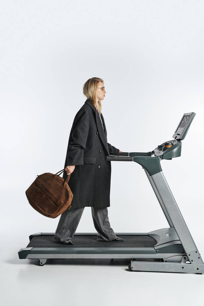 attractive sophisticated woman in fashionable black coat posing on treadmill on gray background - Photo, Image