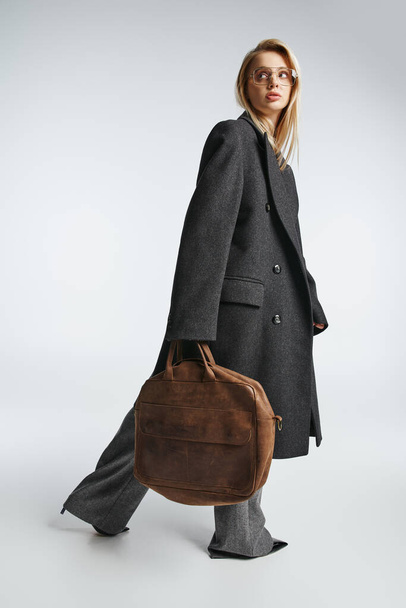 aantrekkelijk sierlijke vrouw met blond haar in zwart stijlvolle jas met bruine tas en wegkijken - Foto, afbeelding