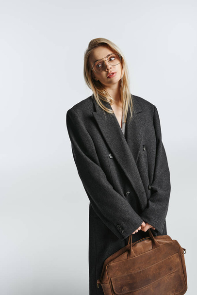 atractiva mujer elegante con el pelo rubio en negro elegante abrigo sosteniendo bolsa marrón y mirando hacia otro lado - Foto, imagen