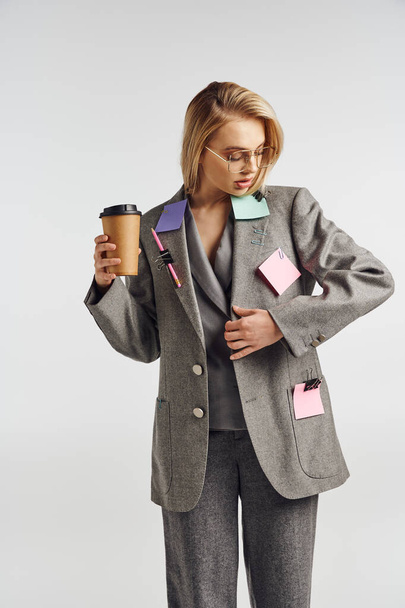 modieuze aantrekkelijke vrouw in grijs pak met briefpapier op poseren met koffie en weg te kijken - Foto, afbeelding
