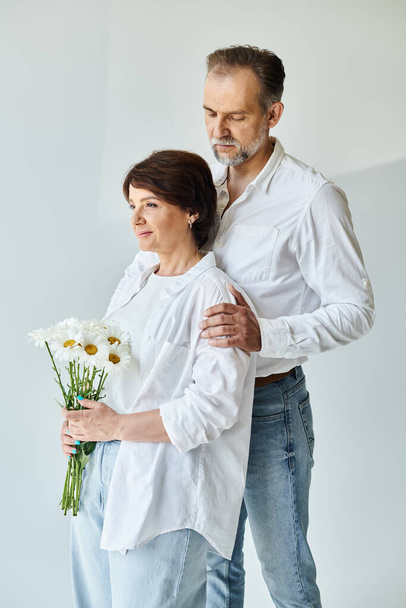 Portrait of mature man in white shirt hugging attractive woman holding flowers on grey background - Foto, afbeelding
