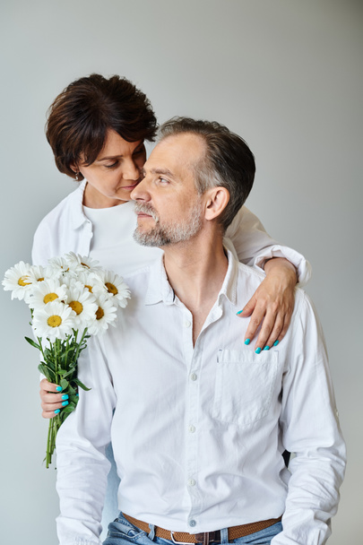 Atractiva mujer sosteniendo flores y abrazando al marido maduro en camisa blanca sobre fondo gris - Foto, imagen