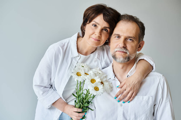 Retrato de pareja de mediana edad con flores sobre fondo gris, mujer abrazándose y apoyándose en el hombre - Foto, Imagen