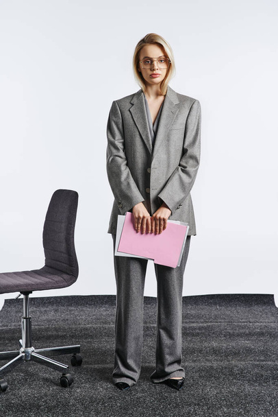 beautiful woman with stylish glasses in gray chic suit posing with paperwork and looking at camera - Photo, Image