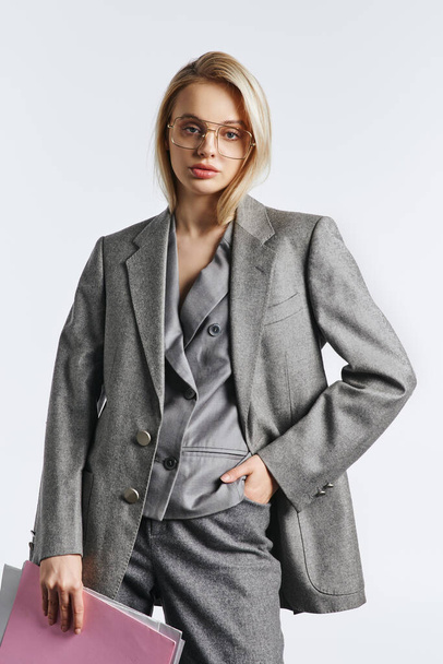 beautiful woman with stylish glasses in gray chic suit posing with paperwork and looking at camera - Zdjęcie, obraz