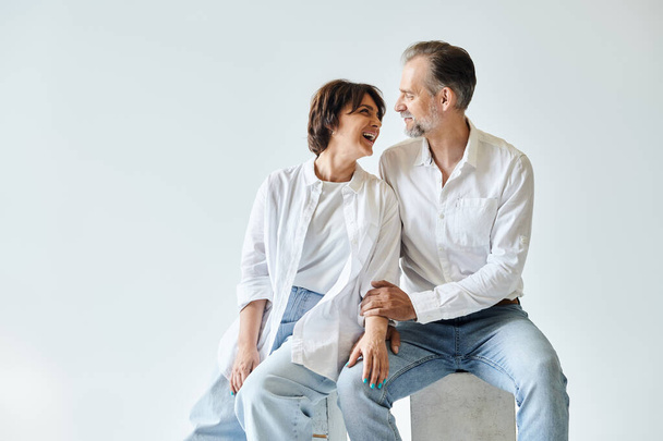 Portrét páru středního věku sedícího a držícího se za ruce na šedém pozadí a dívajícího se jeden na druhého - Fotografie, Obrázek