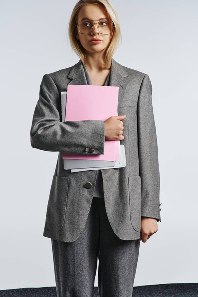 attractive chic woman with stylish glasses in gray suit posing with paperwork and looking away - Foto, imagen