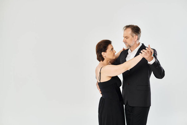 A woman in black dress and man in dark suit embracing on grey background, in tango pose - Foto, imagen