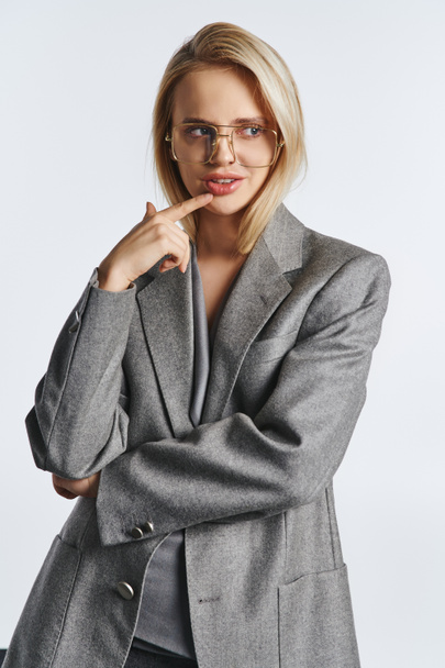veselá atraktivní žena s brýlemi v šedém stylovém obleku pózuje na šedém pozadí a dívá se jinam - Fotografie, Obrázek